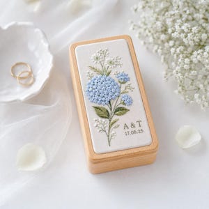May include: A rectangular wooden ring box featuring an embroidered top with light blue hydrangea flowers, green leaves, and white filler flowers. The embroidery includes the text A & T and the date 17.08.25 in a dark grey thread.