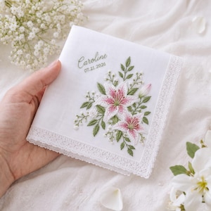 Op de afbeelding: Een witte vierkante zakdoek met geborduurde roze en witte lelies en groene bladeren, afgewerkt met een delicate kanten rand. De stof is gepersonaliseerd met de naam Caroline en de datum 07.21.2026 in groene schrijfletters.