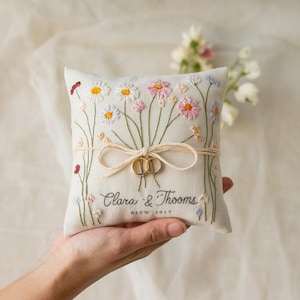 以下が含まれることがあります： 白い、ピンク、紫色のデイジーやその他の花が刺繍された、花の模様のリングピロー。2つのゴールドリングがベージュのリボンで取り付けられています。「Clara & Thoom」と「2017」の文字が刺繍されています。