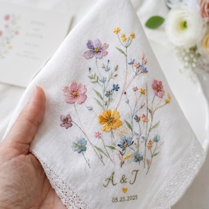 Peut inclure: Un mouchoir en tissu blanc orné d'une broderie florale délicate dans des tons de rose, jaune, violet et bleu. Le motif comprend les initiales brodées A & J, un petit cœur et la date 25.05.2025, avec une bordure en dentelle sur le pourtour.