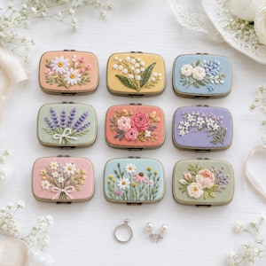 Puede incluir: Nueve joyeros rectangulares de metal con tapas de tela bordada que presentan varios diseños florales, incluyendo margaritas, lavanda y rosas. Debajo de las cajas hay un anillo de plata y un par de pequeños pendientes de perlas blancas.