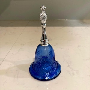 May include: A decorative bell with a cobalt blue glass base and a silver-colored handle. The bell has an ornate, textured design on the glass. The handle is topped with a pineapple-shaped finial. The bell is a decorative item.