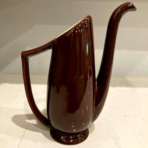 May include: A glossy, dark brown ceramic pitcher with a long, curved spout and a handle. The pitcher has a wide base and a slightly flared opening. The interior is a cream color. The design is reminiscent of a vintage coffee pot.