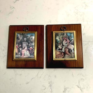 May include: Two framed pictures with gold frames and brown wood backing. The pictures depict scenes of people in a garden setting.