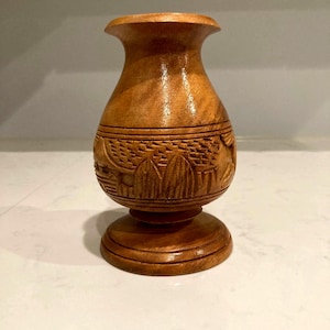 May include: A small, handcrafted wooden vase with a light brown finish. The vase features carved details around the middle and a flared top. The base is round and stable. The vase is approximately 6 inches tall.