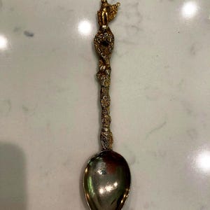 May include: Ornate silver spoon with a detailed gold-colored handle. The handle features a sculpted figure at the top and intricate floral and foliate designs along its length. The spoon's bowl is oval and polished.