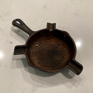 May include: A small, round, dark brown cast iron ashtray shaped like a miniature skillet with a single handle and three integrated cigarette rests. The interior base features embossed text reading Wagner Ware Sidney -O- Foundry Co.