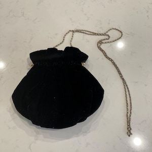 May include: A black velvet drawstring bag with a gold-colored chain strap. The bag has a rounded shape and a gathered top. The chain is delicate and appears to be made of small, linked metal pieces. The bag is on a white surface.