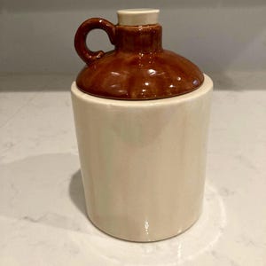 May include: A brown ceramic jar with a white lid and a handle. The jar has a rustic, vintage look.