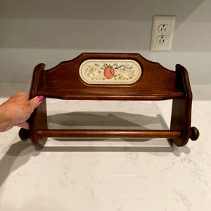 May include: A dark brown wooden paper towel holder featuring a small shelf and a central ceramic tile insert. The tile displays a decorative illustration of two white geese flanking a peach-colored fruit, accented with a blue ribbon and small flowers.