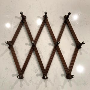 May include: An expandable wall-mounted coat rack featuring a dark brown wooden accordion-style frame with vertical ribbing. The rack includes ten pegs, each topped with a small, round, white ceramic knob, arranged in a diamond-shaped lattice pattern.