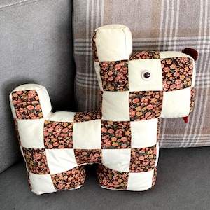 May include: A handmade dog-shaped pillow with a patchwork design. The pillow features alternating squares of cream-colored fabric and fabric with a floral pattern in shades of brown, orange, and yellow. It has a single button eye and a small red pom-pom tail.