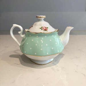 May include: A vintage-style teapot with a mint green polka-dot design. The teapot features a white base and lid, accented with gold trim and a delicate pink rose motif. The handle and spout are white.