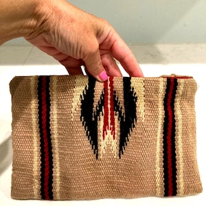 May include: A rectangular woven clutch in shades of tan, black, red, and cream. The clutch features a central geometric design in black and cream, flanked by vertical red stripes. The overall texture appears to be coarse.