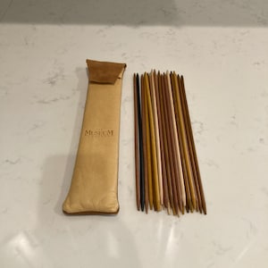 May include: A set of wooden knitting needles in various shades of brown and tan, next to a tan leather case with a brown flap. The case has the text "The Museum" printed on it.