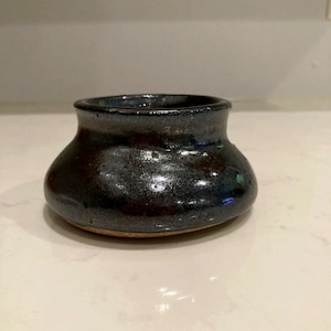 May include: A small, dark ceramic bowl with a wide, rounded body and a narrow opening. The bowl has a glossy, metallic finish with shades of black, blue, and green. The base of the bowl is a lighter, unglazed color.