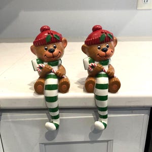 May include: Two brown bear figurines wearing red knit hats with green holly accents. Each bear holds a red and white candy cane and features long, dangling legs with green and white horizontal stripes, designed to sit on a ledge or shelf edge.