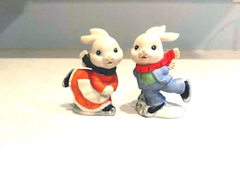 May include: Two ceramic bunny figurines, one in a red and white outfit, the other in blue and green, both wearing ice skates. The bunnies have white fur and are wearing scarves. The figurines are on a white surface.