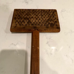 May include: A vintage wooden wool carding tool with a rectangular head covered in metal pins and a long handle. The wood has a warm, brown tone, and the pins are dark. This tool is used for preparing wool fibers for spinning or felting.