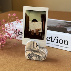 May include: A heart-shaped photo holder with a swirling black and white pattern. A polaroid photo is held in the holder, showing a person reading a book. The book's title is visible. The holder sits on a brown surface.