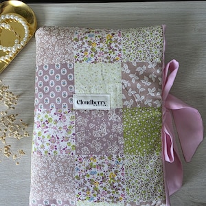 May include: A patchwork journal with a variety of floral and patterned fabrics in shades of purple, green, and white. The journal is tied with a pink satin ribbon. The label reads "Cloudberry Knots".