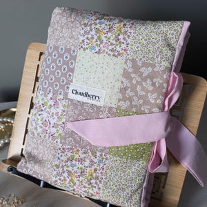 Puede incluir: Una funda de libro de patchwork floral con un lazo rosa, colocada en un atril de madera. La funda presenta varios patrones florales pastel y una etiqueta que dice "Cloudberry Knots".