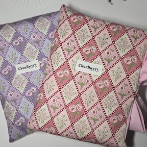 Puede incluir: Dos cojines decorativos con estampados florales. Uno es lavanda y el otro rosa. Ambos cojines tienen un patrón de diamantes con flores y el texto "Cloudberry Knots". Los cojines son un artículo decorativo suave.