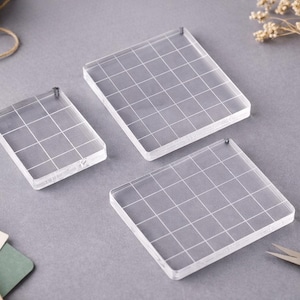May include: Three clear acrylic stamping blocks with a grid pattern, in various sizes, are displayed on a grey surface. Each block has a small metal pin. Other craft supplies, such as twine, scissors, and paper, are also visible.