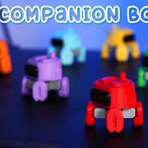 Könnte beinhalten: Eine Sammlung kleiner, farbenfroher Roboter-Spielzeuge in verschiedenen Farben, darunter Rot, Lila, Orange, Blau, Gelb und Grün. Jeder Roboter hat einen rechteckigen schwarzen Bildschirm und ein einzigartiges Design. Der Text "COMPANION BOT" ist zu sehen.