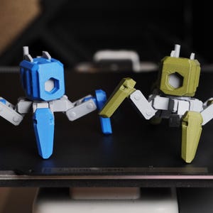 May include: Two 3D-printed robot toys. One is blue and grey, the other is olive green and grey. Each has a cube-shaped head with a hexagonal opening and articulated legs. The toys are on a dark surface.