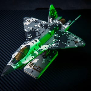Op de afbeelding: Gedetailleerd model van een straaljager met een camouflagepatroon in grijstinten, zwart en wit, geaccentueerd met felgroen. Het model bevat een bijpassende basis met de tekst "SU-57 CHECKMATE".