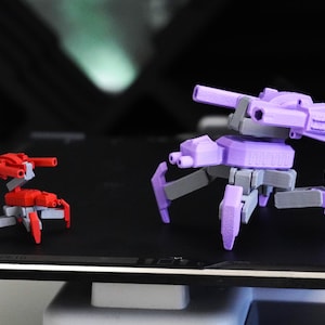 Könnte beinhalten: Zwei 3D-gedruckte Spielzeugroboter. Einer ist rot und grau, der andere ist lila und grau. Beide haben ein Panzer-ähnliches Design mit mehreren Beinen und einer Kanone. Die Spielzeuge befinden sich auf einer schwarzen Oberfläche.