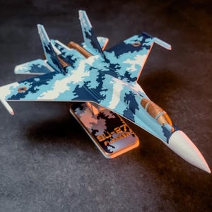 Op de afbeelding: Modelvliegtuig van een SU-27 Flanker straaljager met een blauw-wit camouflagepatroon. Het model staat op een standaard met de tekst "SU-27 Flanker".