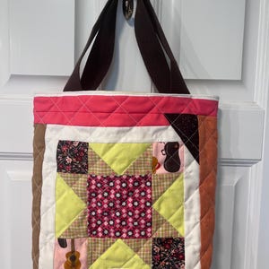Pode incluir: Uma bolsa tote acolchoada com alças marrons. A bolsa é feita de tecido patchwork em tons de rosa, marrom, branco e verde. O tecido apresenta padrões florais e instrumentos musicais.