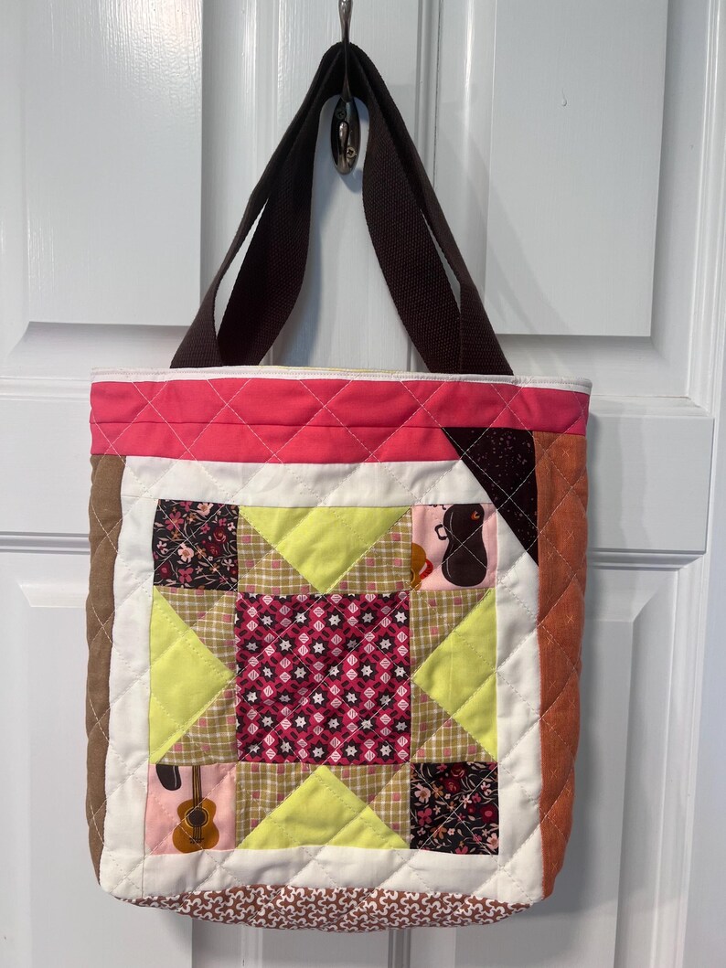 May include: A quilted tote bag with brown straps. The bag is made of patchwork fabric in shades of pink, brown, white, and green. The fabric features floral patterns and musical instruments.