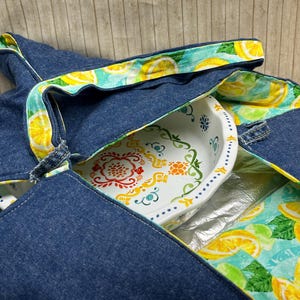 Puede incluir: Un portador de cazuela de mezclilla con un forro estampado de limones coloridos. El portador tiene un asa y está diseñado para contener una fuente para hornear. El plato tiene un fondo blanco con un diseño floral colorido.