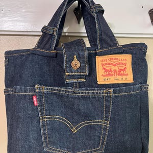 May include: A dark blue denim tote bag featuring two sturdy handles, a central snap button closure, and an exterior pocket. A tan leather-like patch with the text Levi Strauss & Co. 514 18REG 29 29 is attached, along with a small red Levi's tab.