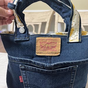 Op de afbeelding: Een handgemaakte tas van donkerblauwe spijkerstof, voorzien van een opvallend bruin Levi Strauss & Co. leren label en een achterzak met een rood Levi's label. De handvatten zijn gevoerd met een witte stof met een patroon van kleine gele bijen.