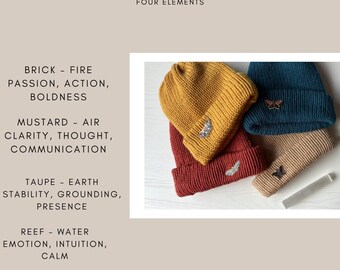 Könnte beinhalten: Vier gestrickte Mützen in verschiedenen Farben, jede mit einem kleinen Schmetterlingsanhänger. Die Mützen sind mit den Elementen Feuer, Luft, Erde und Wasser beschriftet.
