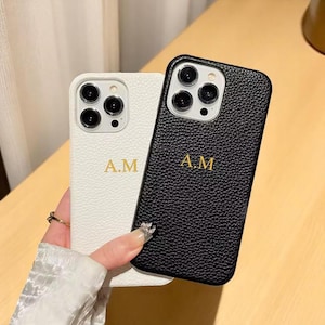 Puede incluir: Se muestran dos fundas de teléfono personalizadas, una blanca y otra negra. Ambas fundas presentan las iniciales "A.M." en letras doradas. La funda blanca tiene una superficie texturizada, mientras que la funda negra tiene una textura similar al cuero. Las fundas están diseñadas para un smartphone.