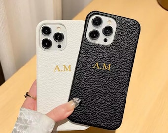 May include: Two personalised phone cases are shown, one white and one black. Both cases have the initials "A.M." in gold lettering. The white case has a textured surface, while the black case has a leather-like texture. The cases are designed for a smartphone.