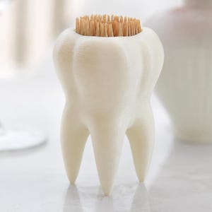 Puede incluir: Un palillero blanco con forma de diente, lleno de palillos de madera. El soporte tiene un diseño detallado y realista, con tres raíces en la base. El fondo incluye una copa de vino y un jarrón blanco, lo que sugiere un comedor o una cocina.