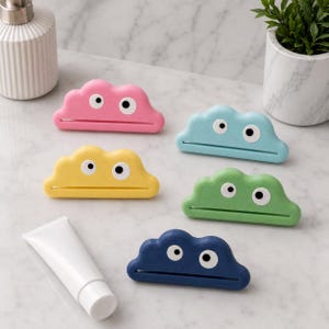 Könnte beinhalten: Eine Sammlung von wolkenförmigen Tubenquetschern in Rosa, Gelb, Blau, Grün und Marineblau. Jeder hat zwei Kulleraugen. Eine weiße Tube Produkt ist ebenfalls vorhanden. Diese sind dazu bestimmt, den Inhalt von Tuben herauszudrücken.