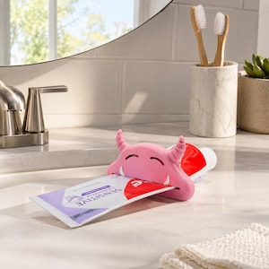 Può includere: Uno spremi-tubo per dentifricio a forma di mostro rosa con un tubetto di dentifricio rosso e bianco. Il tubetto è etichettato "SENSITIVE". Lo spremi-tubo ha corna ricurve e una faccia sorridente.