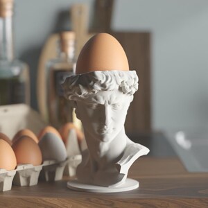 May include: A white matte finish egg cup shaped like a classical male bust sculpture. The head of the bust features detailed hair and facial features, with a smooth circular base. A single brown egg is placed securely in the top of the head.