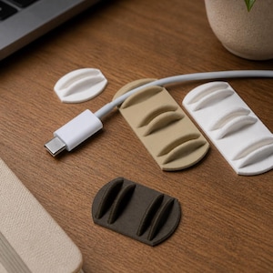 May include: Cable tidies in white, beige, and dark brown, with curved slots for holding cords. A white USB-C cable is connected to a laptop. A small potted plant and a notebook are also on the wooden surface.