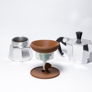Puede incluir: Un soporte marrón para cafetera, con una base redonda y una parte superior en forma de cuenco, colocado entre dos cafeteras de espresso plateadas. El soporte está diseñado para sostener un filtro de café. El fondo es blanco liso.