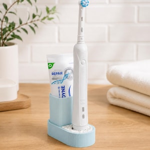 May include: A white electric toothbrush with a blue and white brush head is in a light blue holder next to a tube of toothpaste. The toothpaste tube has the word "DYNE" printed on it. The holder is on a wooden surface.