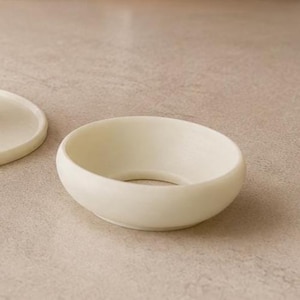 Peut inclure: Bol en c&eacute;ramique blanc cass&eacute; de forme arrondie avec une ouverture circulaire au centre. Le bol a une finition lisse et mate et repose sur une surface de ton neutre. Le bol fait partie d'un ensemble.