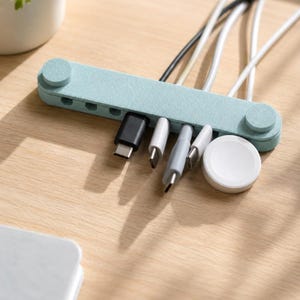 May include: A light blue cable organiser bar with a textured finish, featuring circular end caps and multiple slots holding various charging cables with USB-C connectors, alongside a round white magnetic charging puck for a smartwatch.